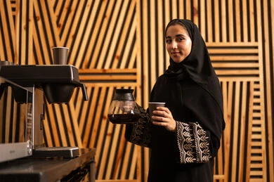 Women's professions and jobs, working in the administrative and office sector, break room, an Arab Gulf Saudi woman wearing an abaya and hijab using a quick coffee maker, a local Saudi national product, enjoying preparing fresh hot beverages.