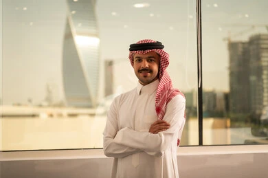 The evolution in the design of offices and upscale companies, working in the administrative and office sector, professions and jobs in Saudi Arabia, an Arab Gulf Saudi man wearing traditional attire and a ghutrah stands next to glass windows overlooking the towers and skyscrapers, looking at the camera happily.