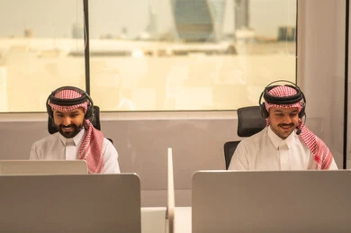 Designated spaces for each employee in the company, focus and meticulous work, dialogues and discussions within the team, business and company development, work meetings and project completion, discussing plans and explaining work tasks, Arab Saudi Gulf colleagues wearing traditional attire and shemagh, using headphones and laptops.