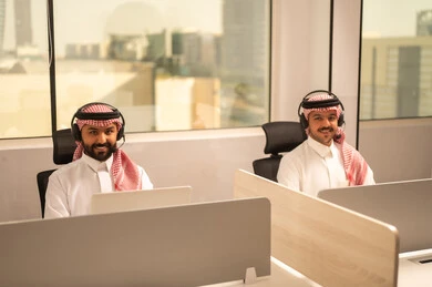 Designated spaces for each employee in the company, focus and meticulous work, dialogues and discussions within the team, business and company development, work meetings and project completion, discussing plans and explaining work tasks, Arab Saudi Gulf colleagues wearing traditional attire and shemagh, using headphones and laptops.