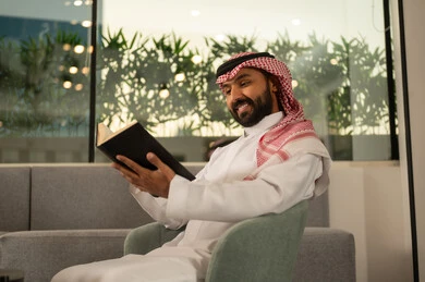 Reading books attentively, enjoying leisure time, the culture of self-learning, a smiling Arab Gulf Saudi man wearing traditional attire and a ghutrah holding an educational book in his hand, gestures of focus and engagement, reading novels and stories.