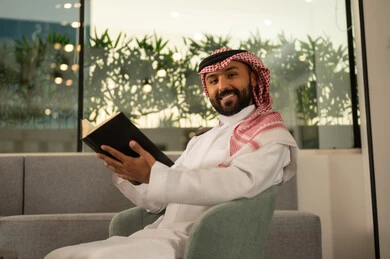 الاستمتاع باوقات الفراغ ، ثقافة التعلم الذاتي، قراءة الكتب بتمعن ، رجل عربي خليجي سعودي مبتسم يرتدي الثوب التقليدي والشماغ يحمل بيده كتاب تثقيفي ، إيماءات التركيز والاندماج ، قراءة الروايات والقصص .