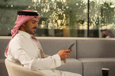 رجل عربي خليجي سعودي مبتسم يرتدي الثوب التقليدي والشماغ يقوم باستخدام الهاتف المحمول داخل مقر الشركة، التواصل مع الاهل والاصدقاء، ايماءات الفرح والسرور، دمج التقنية والتكنولوجيا في الحياة اليومية والعملية