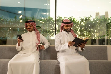Enjoying hot drinks, reading and a love for literature, Arab Gulf Saudi employees sitting inside the office, working in high-end Saudi companies, working on mobile phones, discussing projects and following up on work strategies.
