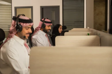 Designated spaces for each employee in the company, focus and meticulous work, dialogues and discussions within the team, business and company development, work meetings and project completion, discussing plans and explaining work tasks, Arab Saudi Gulf colleagues wearing traditional attire and shemagh, using headphones and laptops.