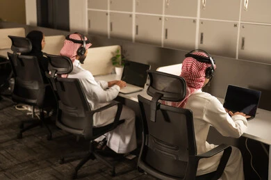 Discussion of plans and explanation of work tasks, work meetings and project completion, business and company development, designated spaces for each employee in the company, focus and meticulous work, dialogues and discussions within the team, Arab Saudi Gulf colleagues wearing traditional attire and shemagh, using headphones and laptops.