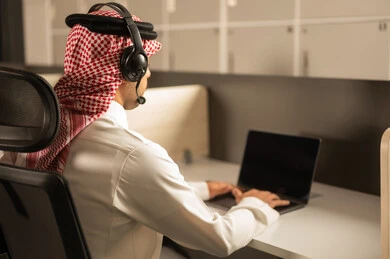 اجتماع عمل وانجاز المشاريع, أماكن مخصصة لكل موظف في الشركة، التركيز والعمل المتقن, الحوارات والنقاشات ضمن الفريق,تطوير الاعمال والشركات,مناقشة الخطط وشرح مهام الاعمال, موظف عربي خليجي سعودي يرتدي الزى التقليدي والشماغ وسماعات الرأس يستخدم الحاسوب المحمول
