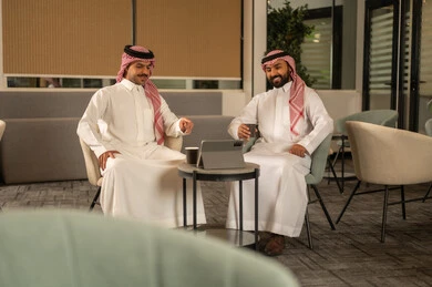 Enjoying reading books, two Saudi Gulf Arab employees sitting inside the office, working in prestigious Saudi companies, working on a tablet, discussing projects and following up on the work strategy.