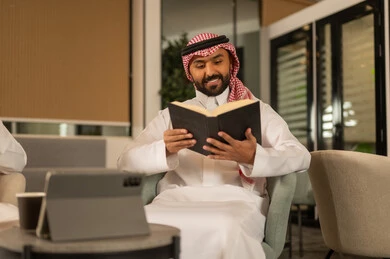 ثقافة التعلم الذاتي، الاستمتاع باوقات الفراغ ، قراءة الكتب بتمعن ، رجل عربي خليجي سعودي مبتسم يرتدي الثوب التقليدي والشماغ يحمل بيده كتاب تثقيفي ، إيماءات التركيز والاندماج ، قراءة الروايات والقصص .