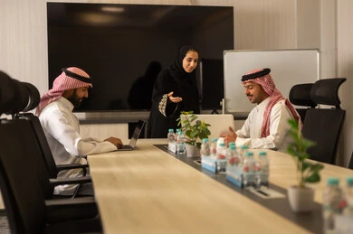 Collaboration and understanding among the team, exchanging experiences and successful agreements, organizing and managing work objectives, a comfortable work environment, Saudi employees using laptops, happily explaining the required tasks, Arab Saudi Gulf colleagues in a meeting to study the project and objectives in detail.