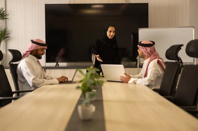 Collaboration and understanding among the team, exchanging experiences and successful agreements, organizing and managing work objectives, a comfortable work environment, Saudi employees using laptops, happily explaining the required tasks, Arab Saudi Gulf colleagues in a meeting to study the project and objectives in detail.
