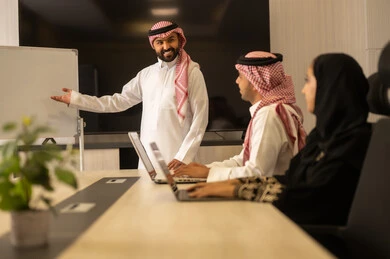 Organizing and managing work objectives, a comfortable work environment, cooperation and understanding among the team, exchanging experiences and successful agreements, Saudi employees using a laptop, happily explaining the required tasks, Arab Saudi Gulf colleagues in a meeting to study the project and objectives in detail.