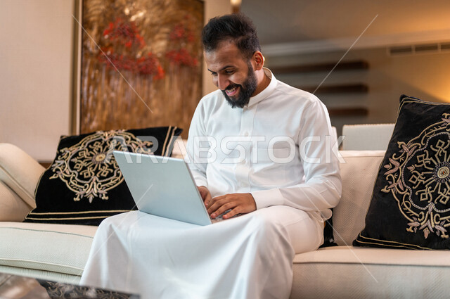 رجل عربي خليجي سعودي يجلس في غرفة المعيشة ويتصفح الانترنت من خلال الحاسوب المحمول، استخدام اللابتوب في انجاز الاعمال، قضاء وقت ممتع، العمل عن بعد