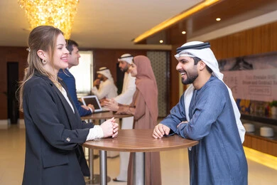 Organization and strategic planning to achieve goals, networking and building social relationships, a young Arab Gulf Emirati discussing work methods and professional development with a foreign businesswoman, concluding new deals, holding a meeting inside the company headquarters, teamwork and team spirit, background of businessmen.