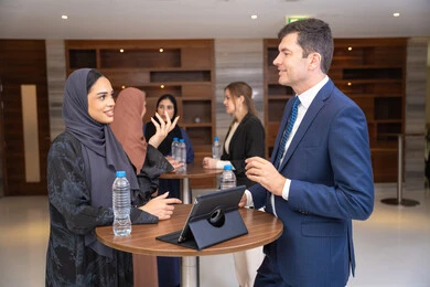 Integrating technology with daily tasks, closing new deals, strategic planning and organization to achieve goals, networking and building social relationships, an Arab Gulf Emirati woman discusses work methods and professional development with a foreign businessman, holding a meeting inside the company headquarters, background of businessmen.