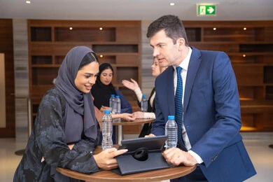 استخدام التابلت مع المهام اليومية، عقد الصفقات الجديدة ،التخطيط الاستراتيجي والتنظيم لتحقيق الأهداف، التعارف وبناء العلاقات الاجتماعية، سيدة عربية خليجية اماراتية تناقش اساليب العمل والتطوير المهني مع سيد اعمال اجنبية، عقد اجتماع داخل مقر الشركة، خلفية رجال اعمال