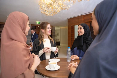 Drinking hot beverages during work, discussion and understanding among employees, exchanging experiences and information to achieve goals, strategic planning and organization to achieve objectives, concluding new deals, a group of Arab Gulf Emirati women discussing work methods and professional development with a foreign businesswoman.
