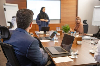 Integrating technology into work, professions and jobs for women, using a display screen for clearer explanations, ensuring a clear and comprehensive understanding of the work, discussion and understanding among employees, strategic plans to achieve goals, an Arab Gulf Emirati woman explaining work methods and professional development to a group of Arab and foreign individuals.