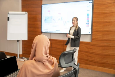 Exchange of experiences between different companies, dialogue and understanding among employees, unity and teamwork, office professions and jobs, using a display screen for clearer explanations, ensuring a clear and comprehensive understanding of the work, strategic plans to achieve goals, a foreign businessman discussing work methods and professional development within an Emirati company.