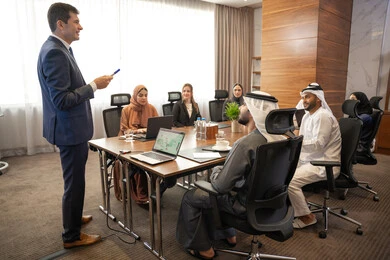 Employee development and training, clarifying the company's goals and policies, dialogue and understanding among employees, unity and teamwork, office professions and jobs, using a display screen for clearer explanations, ensuring a clear and comprehensive understanding of the work, a foreign businessman discussing work methods and professional development within an Emirati company.