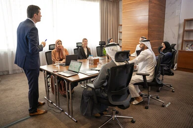 Skills of presentation in a business meeting, dialogue and understanding among employees, unity and teamwork, office professions and jobs, using a projector for clearer explanations, ensuring a clear and comprehensive understanding of the work, strategic plans to achieve goals, a foreign businessman discussing work methods and professional development within an Emirati company.