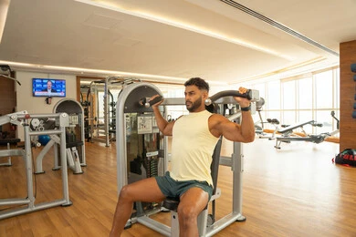 Training with determination to maintain a healthy lifestyle; the importance of exercise for building a strong body; increasing flexibility and strength; an Emirati Gulf Arab man wearing sportswear and exercising on a shoulder exercise machine; building muscle mass; strengthening muscles and nerves.