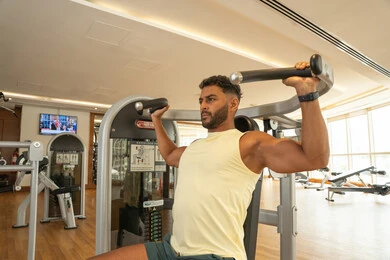 أنشطة ورياضات لتقوية العضلات, نشاط شبابي يساعد على اللياقة البدنية, أهمية الرياضة لبناء جسد قوي، رجل عربي خليجي إماراتي يرتدي الملابس الرياضية و يتمرن على آلة تمرين الأكتاف, اتباع أسلوب حياة صحي, ايماءات القوة والثقة