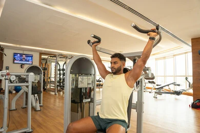A youth activity that promotes physical fitness; the importance of sports for building a strong body; an Emirati Gulf Arab man wearing sportswear and exercising on a shoulder exercise machine; adopting a healthy lifestyle; maintaining body balance; gestures of strength and confidence.