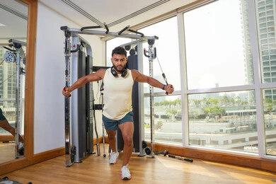 أهمية الرياضة لبناء جسد قوي، زيادة المرونة و القوة, رجل عربي خليجي إماراتي يرتدي الملابس الرياضية يتمرن على جهاز الكابل و يضع سماعه الرأس حول رقبته, اتباع أسلوب حياة صحي, استخدام الأجهزة الرياضية