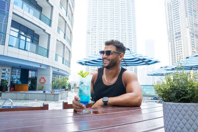 مفهوم الوسامة والثقة بالنفس، استخدام النظارات الشمسية السوداء لحماية العيون من أشعة الشمس،  الاسترخاء في الهواء الطلق, رجل عربي خليجي اماراتي يرتدي الزي الكاجوال و يقوم بشرب مشروب طازج, الاسترخاء في الهواء الطلق