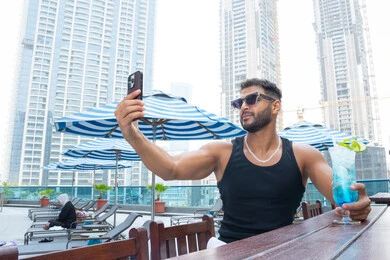 مفهوم الأناقة والرجولة، الهروب من حرارة الصيف باحتساء المشروبات الباردة، الأجواء الممتعة الهادئة, مفهوم الاستجمام والاختلاء, الاسترخاء في الهواء الطلق, رجل عربي خليجي اماراتي يرتدي الزي الكاجوال و النظارة الشمسية و يقوم بالتقاط صور سيلفي للتذكار