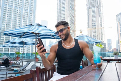 Wearing fashionable sunglass, escaping the summer heat with cold drinks, enjoying a pleasant and tranquil atmosphere, the concept of relaxation and seclusion, unwinding outdoors, an Emirati Gulf Arab man wearing casual clothes and browsing his mobile phone.