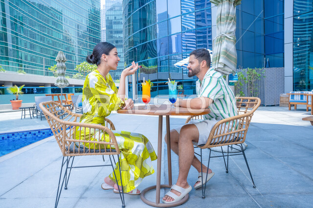 Gestures of joy and camaraderie, family-friendly recreational activities, trying to alleviate the summer heat with refreshing drinks, relaxing outdoors, having fun, engaging in enjoyable holiday activities, an Emirati Gulf Arab couple in casual attire sitting in a resort in the UAE