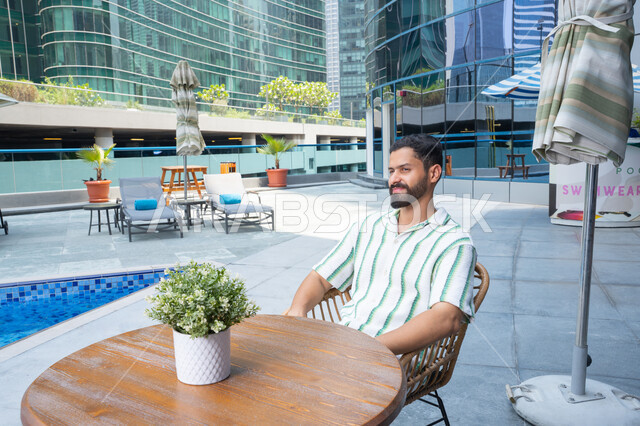 مفهوم الراحة والاسترخاء, الاستمتاع بالأجواء الهادئة, رجل عربي خليجي اماراتي مبتسم يرتدي الزي الكاجوال و يجلس بجانب المسبح, القيام بالأنشطة الممتعة في الإجازة, قضاء الأوقات المسلية, ايماءات السرور و السعادة