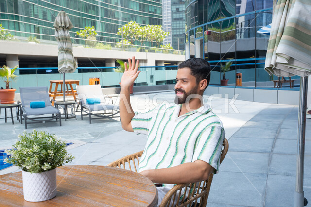 Greetings and salutations, enjoying a peaceful atmosphere outdoors, escaping the summer heat with cold drinks, the concept of relaxation and solitude, unwinding in the open air, a smiling Emirati Gulf Arab man in casual attire