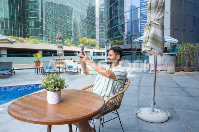 Going on a leisure trip, escaping the summer heat, the concept of relaxation and seclusion, unwinding outdoors, an Emirati Gulf Arab man wearing casual clothes and taking selfies as a souvenir, engaging in fun activities during vacation