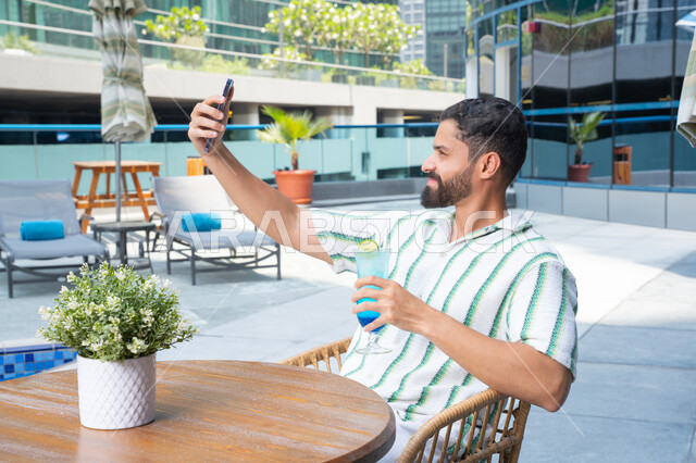 الهروب من حرارة الصيف باحتساء المشروبات الباردة، الأجواء الممتعة الهادئة, مفهوم الاستجمام والاختلاء,  الاسترخاء في الهواء الطلق, رجل عربي خليجي اماراتي يرتدي الزي الكاجوال و يقوم بالتقاط صور سيلفي للتذكار, القيام بالأنشطة الممتعة في الإجازة