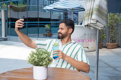 The pleasant, tranquil atmosphere, the concept of relaxation and seclusion, enjoying refreshing cold drinks, relaxing outdoors, an Emirati Gulf Arab man wearing casual clothes taking souvenir selfies, engaging in fun holiday activities