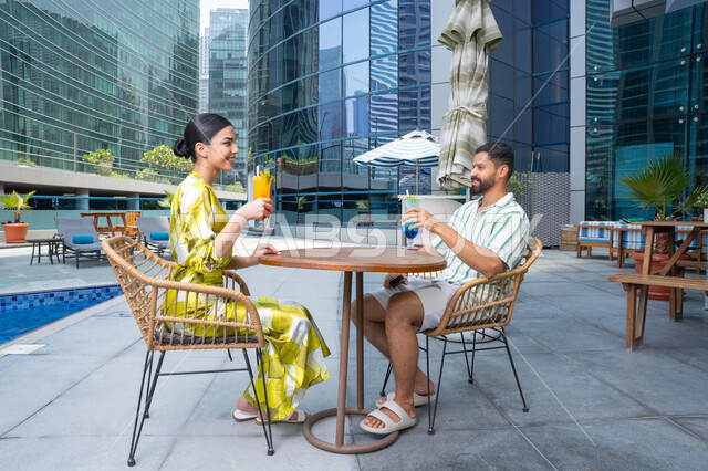 The intimacy and affection between the couple, a happy family atmosphere, the concept of talking and discussing the news, an Emirati Gulf Arab couple wearing casual clothes sitting in one of the resorts in the Emirates and enjoying fresh, refreshing drinks.