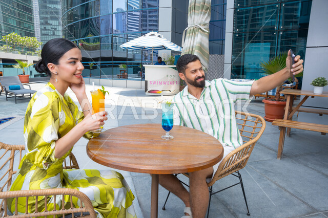 Documenting family moments, enjoying taking memorable selfies on their mobile phones, engaging in fun recreational activities, an Emirati Gulf Arab couple in casual attire sitting in a resort in the UAE and enjoying cold drinks