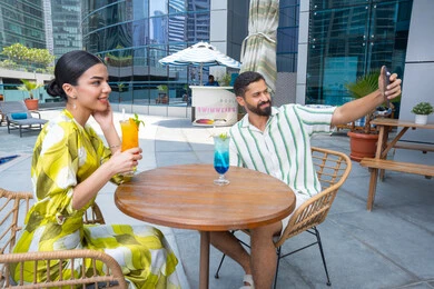 توثيق اللحظات العائلية, الاستمتاع بالتقاط صور سيلفي تذكارية عبر الجوال,  القيام بالأنشطة الترفيهية الممتعة, زوجان عربيان خليجيان إماراتيان يرتديان الزي الكاجوال يجلسان في إحدى المنتجعات في الإمارات و يتناولان المشروبات الباردة