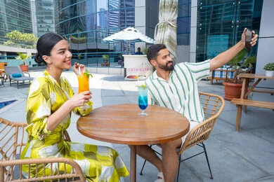 الخروج في الهواء الطلق في وقت النهار, الاستمتاع بالتقاط صور سيلفي تذكارية عبر الجوال,  القيام بالأنشطة الترفيهية الممتعة, زوجان عربيان خليجيان إماراتيان يرتديان الزي الكاجوال يجلسان في إحدى المنتجعات في الإمارات و يتناولان المشروبات الباردة