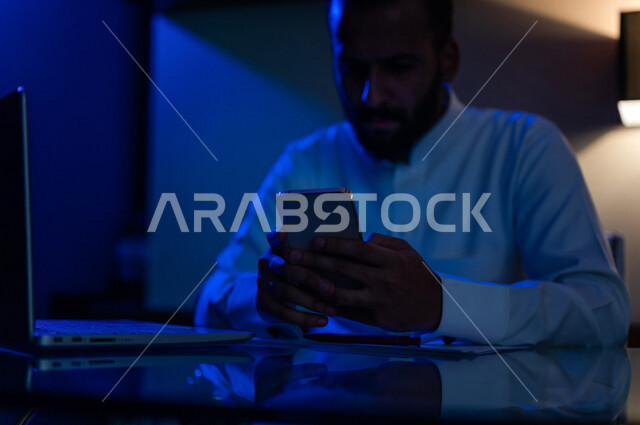 رجل عربي خليجي سعودي يجلس امام الحاسوب ويمسك بيده هاتف محمول، التواصل الاجتماعي، محادثات ودردشة، العمل اونلاين، فريلانسر، اعمال مكتبية