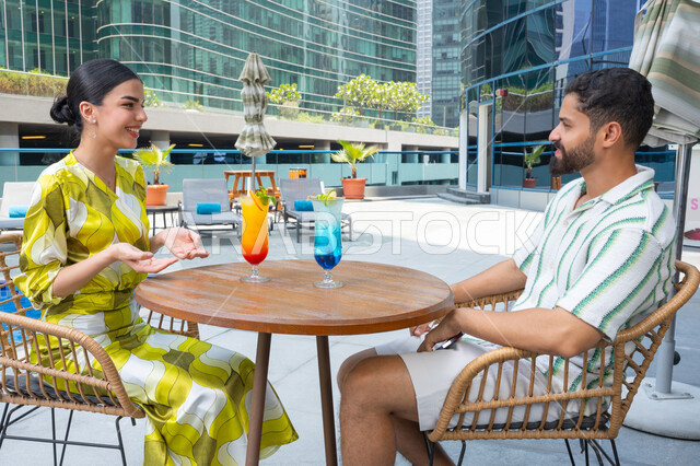 Chatting, relaxing outdoors, enjoying fun family time, engaging in enjoyable leisure activities during vacation: an Emirati Gulf Arab couple, dressed casually, sitting in a resort in the UAE and sipping fresh drinks.