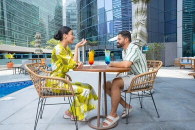 عطلة نهاية الاسبوع, حوارات ونقاشات على الطاولة, الإستجمام في الهواء الطلق, قضاء الأوقات العائلية المسلية, القيام بالأنشطة الترفيهية الممتعة في الإجازة, زوجان عربيان خليجيان إماراتيان يرتديان الزي الكاجوال يجلسان في إحدى المنتجعات في الإمارات و يتناولان المشروبات الباردة