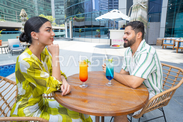 محاولة التخفيف من حرارة الصيف بالمشروبات المنعشة، الإستجمام في الهواء الطلق, قضاء الأوقات العائلية المسلية, القيام بالأنشطة الترفيهية الممتعة في الإجازة, زوجان عربيان خليجيان إماراتيان يرتديان الزي الكاجوال يجلسان في إحدى المنتجعات في الإمارات