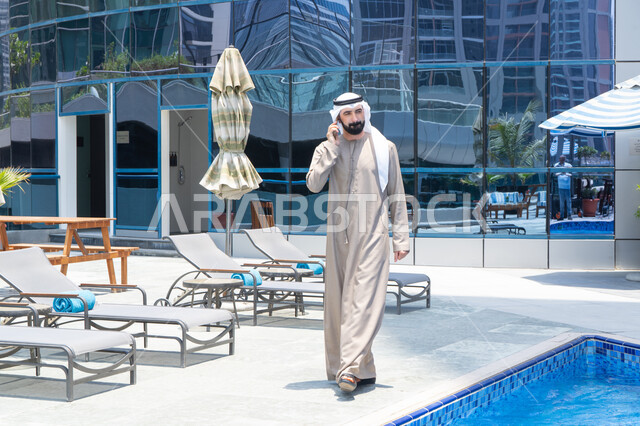 التحدث عبر الهاتف المحمول، التواصل عن بعد, الرد على مكالمة هاتفية عبر الجوال، قضاء اوقات ممتعة بتبادل الاخبار والأحاديث، الإستجمام في الهواء الطلق, المشي على حدود البركة المائية, رجل عربي خليجي اماراتي يرتدي الكندورة والغترة و يقوم بالتنزه