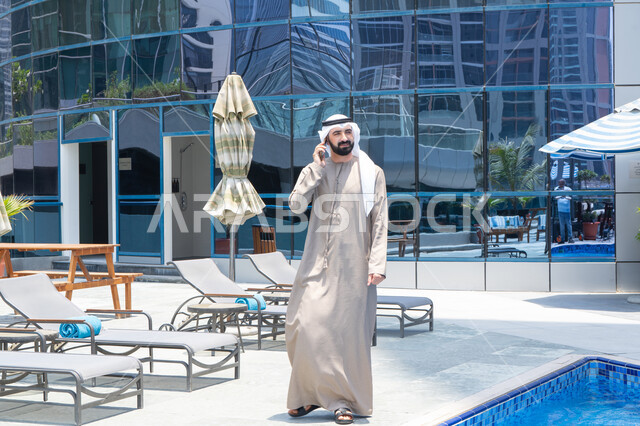 الاطمئنان على الأصدقاء,  التواصل عن بعد, الرد على مكالمة هاتفية عبر الجوال، قضاء اوقات ممتعة بتبادل الاخبار والأحاديث، الإستجمام في الهواء الطلق, المشي على حدود البركة المائية, رجل عربي خليجي اماراتي يرتدي الكندورة والغترة و يقوم بالتنزه