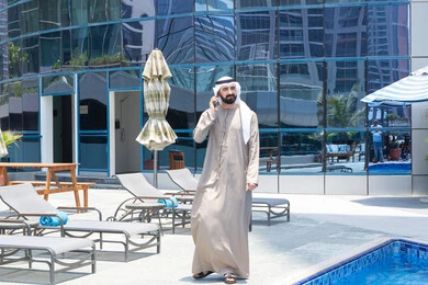 الاطمئنان على الأصدقاء,  التواصل عن بعد, الرد على مكالمة هاتفية عبر الجوال، قضاء اوقات ممتعة بتبادل الاخبار والأحاديث، الإستجمام في الهواء الطلق, المشي على حدود البركة المائية, رجل عربي خليجي اماراتي يرتدي الكندورة والغترة و يقوم بالتنزه