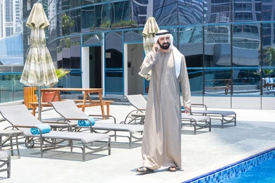 استخدام جهاز تقني حديث ومتطور، الرد على مكالمة هاتفية عبر الجوال، قضاء اوقات ممتعة بتبادل الاخبار والأحاديث، الإستجمام في الهواء الطلق, المشي على حدود البركة المائية, رجل عربي خليجي اماراتي يرتدي الكندورة والغترة و يقوم بالتنزه